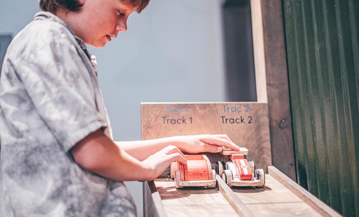 A teenage boy holds two model cars at the top of a wooden, sloped track